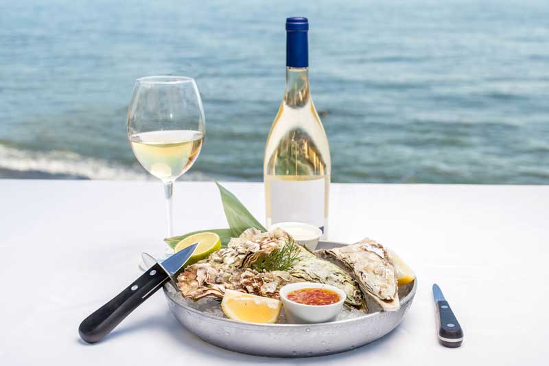 Oysters and a bottle of wine on a white table overlooking the water.